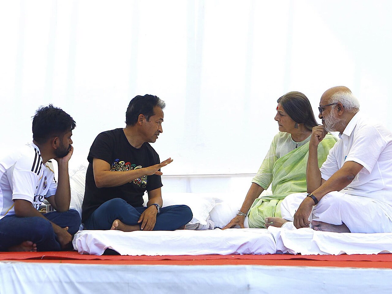 Sonam Wangchuk, cross-legged on a white mattress under a tent, in conversation with CPI(M) leaders Brinda Karat and M. A. Baby seated beside him