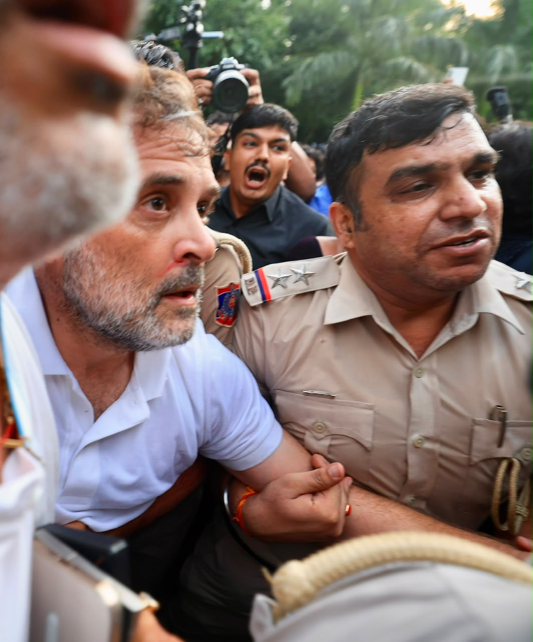 Rahul Gandhi in a white polo shirt, held by the arm by a Delhi Police officer amid a crush of people and press cameras